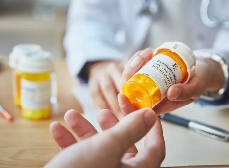 Medical professional handing a prescription bottle to a patient over a table with prescription bottles and pens in the background